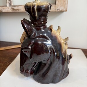 May include: A ceramic horse head decanter with a crown-shaped stopper. The decanter is primarily dark brown with lighter brown and tan accents. The horse's head is detailed, and the crown adds a regal touch.
