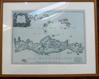 May include: A framed historical map of the island of Candie, featuring detailed topography and nautical labels in French. The map is printed in black ink on a light blue background, enclosed in a light wood frame with a white mat border.