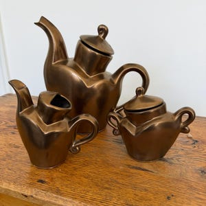 May include: A bronze-colored ceramic tea set, including a teapot, creamer, and sugar bowl, is displayed on a wooden surface. The teapot has a curved spout and handle, and the sugar bowl has a lid. The set has a unique, modern design.
