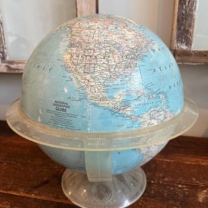 Pode incluir: Um globo terrestre vintage da National Geographic com um oceano azul claro e massas terrestres detalhadas. O globo assenta sobre uma base de plástico transparente com um anel meridiano. O texto "NATIONAL GEOGRAPHIC GLOBE" é visível.