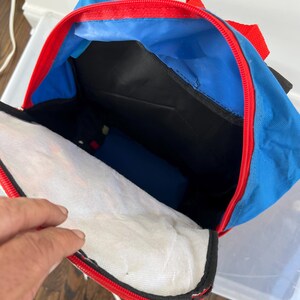 May include: Blue backpack with red trim and a black interior. The backpack is open, revealing a white mesh pocket and a blue interior lining. The zipper is red, and the straps are also red.
