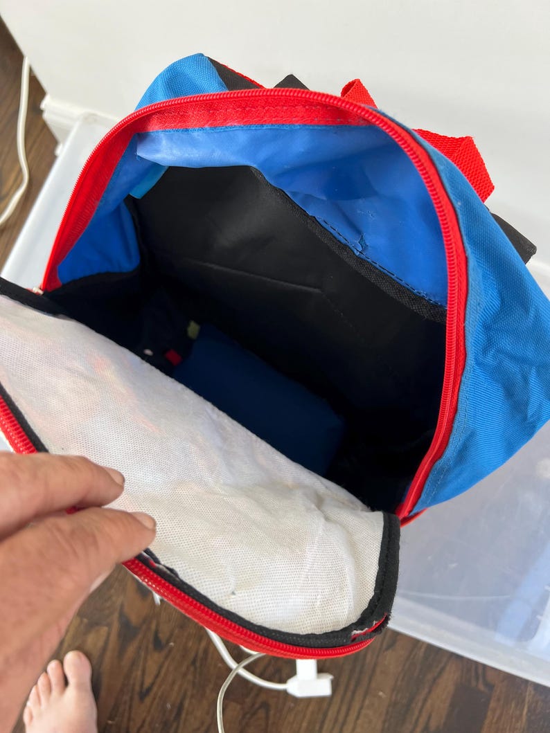 May include: Blue backpack with red trim and a black interior. The backpack is open, revealing a white mesh pocket and a blue interior lining. The zipper is red, and the straps are also red.