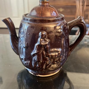 May include: A brown ceramic teapot with a lid and handle. The teapot features a relief design of figures and a scene. The teapot is sitting on a reflective surface.
