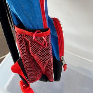 May include: Blue backpack with red accents, a red mesh side pocket, and a red zipper. The backpack has a black strap and a red carabiner clip. The design includes white trim and a red buckle.