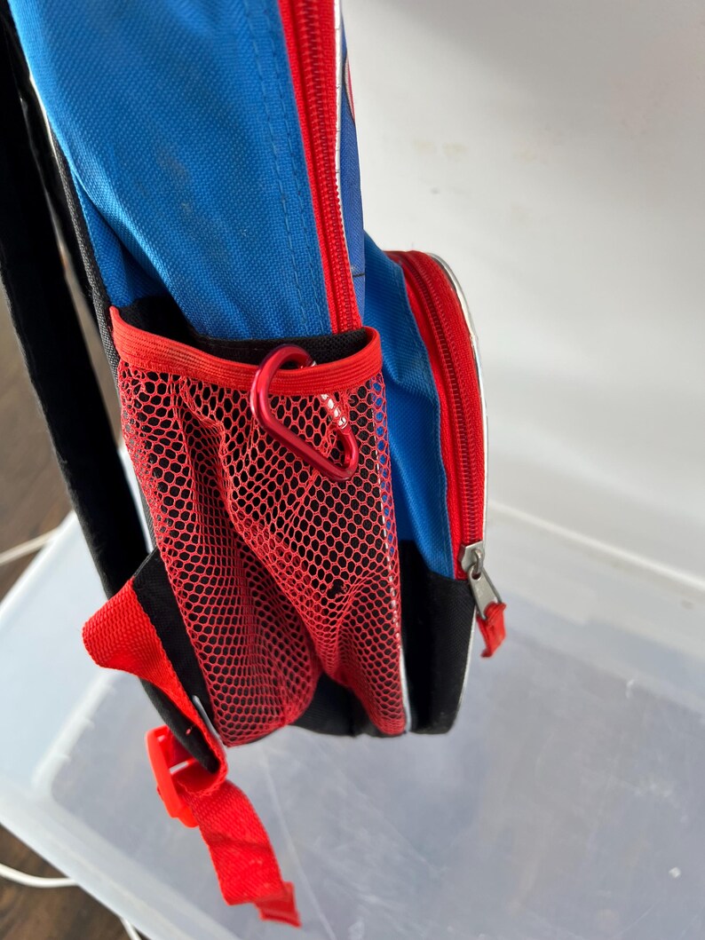 May include: Blue backpack with red accents, a red mesh side pocket, and a red zipper. The backpack has a black strap and a red carabiner clip. The design includes white trim and a red buckle.