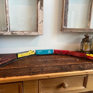 May include: A model train set with a maroon engine, yellow and blue cars, and red boxcars on a curved track. The train cars have text that reads "The Rock", "Delaware Hudson", and "Rock Island". The train set is displayed on a wooden surface.