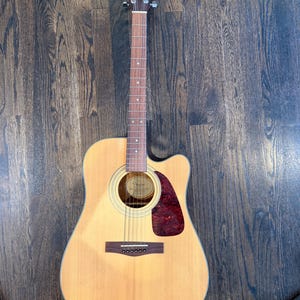 Pu&ograve; includere: Chitarra acustica con finitura in legno naturale. La chitarra presenta un battipenna rosso scuro, una buca e sei corde. La paletta ha meccaniche e il marchio "Fender". La chitarra &egrave; appoggiata su un pavimento in legno scuro.