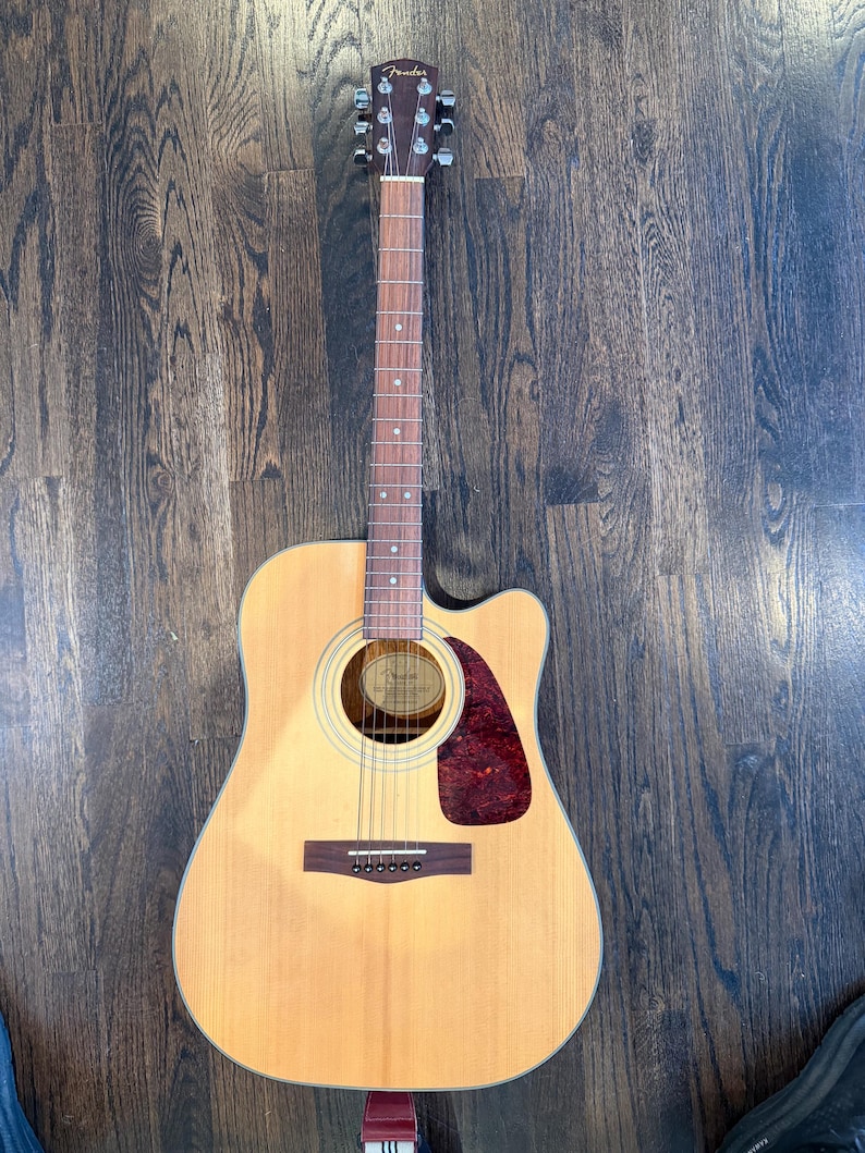 Pu&ograve; includere: Chitarra acustica con finitura in legno naturale. La chitarra presenta un battipenna rosso scuro, una buca e sei corde. La paletta ha meccaniche e il marchio "Fender". La chitarra &egrave; appoggiata su un pavimento in legno scuro.