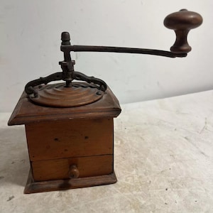 May include: A vintage wooden coffee grinder with a hand crank. The grinder has a square base with a drawer and a metal mechanism on top. The handle and knob are made of wood. The overall color is brown.