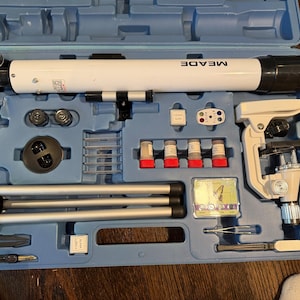 May include: A blue case containing a white telescope with "MEADE" printed on it, a microscope, a tripod, and various scientific tools. The case is open, revealing the organized contents.