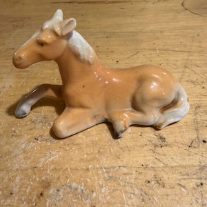 Puede incluir: Figura de caballo de cerámica en reposo. El caballo es principalmente de color marrón claro con crin y cola blancas. La figura tiene un acabado brillante y está sobre una superficie de madera.