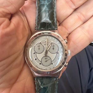 Op de afbeelding: Een Swatch chronograaf horloge met een zilverkleurige kast en een lichtgrijze wijzerplaat. Het horloge heeft drie sub-wijzerplaten en een teal leren band. Het horloge wordt in een hand gehouden.