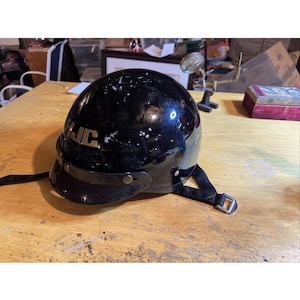 Pode incluir: Capacete de motocicleta preto com viseira e cinta queixeira. O capacete tem as letras "HJC" em branco na lateral. O capacete está sobre uma superfície de madeira.