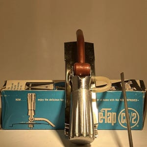 Puede incluir: Un sistema de grifo de cerveza e-Tap CV2 vintage. El grifo tiene un cuerpo plateado con un mango marrón y está montado en una placa de metal. La caja dice "enjoy the delicious fun" y "e-Tap CV2".
