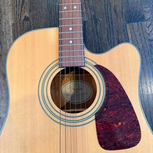 Pu&ograve; includere: Chitarra acustica con finitura in legno naturale. La chitarra presenta una buca rotonda, una tastiera marrone scuro e un battipenna rosso tartarugato. Le corde sono visibili e la paletta &egrave; fuori campo.