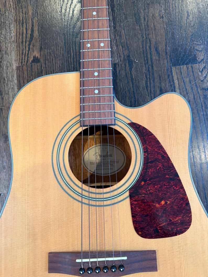 Pu&ograve; includere: Chitarra acustica con finitura in legno naturale. La chitarra presenta una buca rotonda, una tastiera marrone scuro e un battipenna rosso tartarugato. Le corde sono visibili e la paletta &egrave; fuori campo.