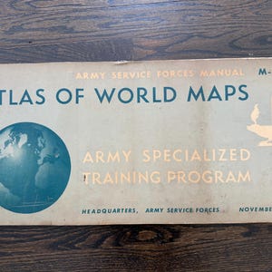 May include: Vintage "Atlas of World Maps" manual from the Army Service Forces, M-101. The cover features a globe illustration and text in teal and gold. Published by the Army Specialized Training Program in November 1943.