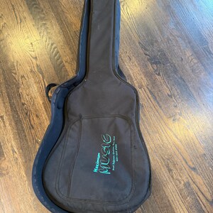 Pu&ograve; includere: Custodia per chitarra nera con chiusura a zip e tasca frontale. La custodia presenta la scritta "Maximum Music" in lettere turchesi. La custodia &egrave; aperta, rivelando un interno blu scuro. La custodia &egrave; su un pavimento in legno.