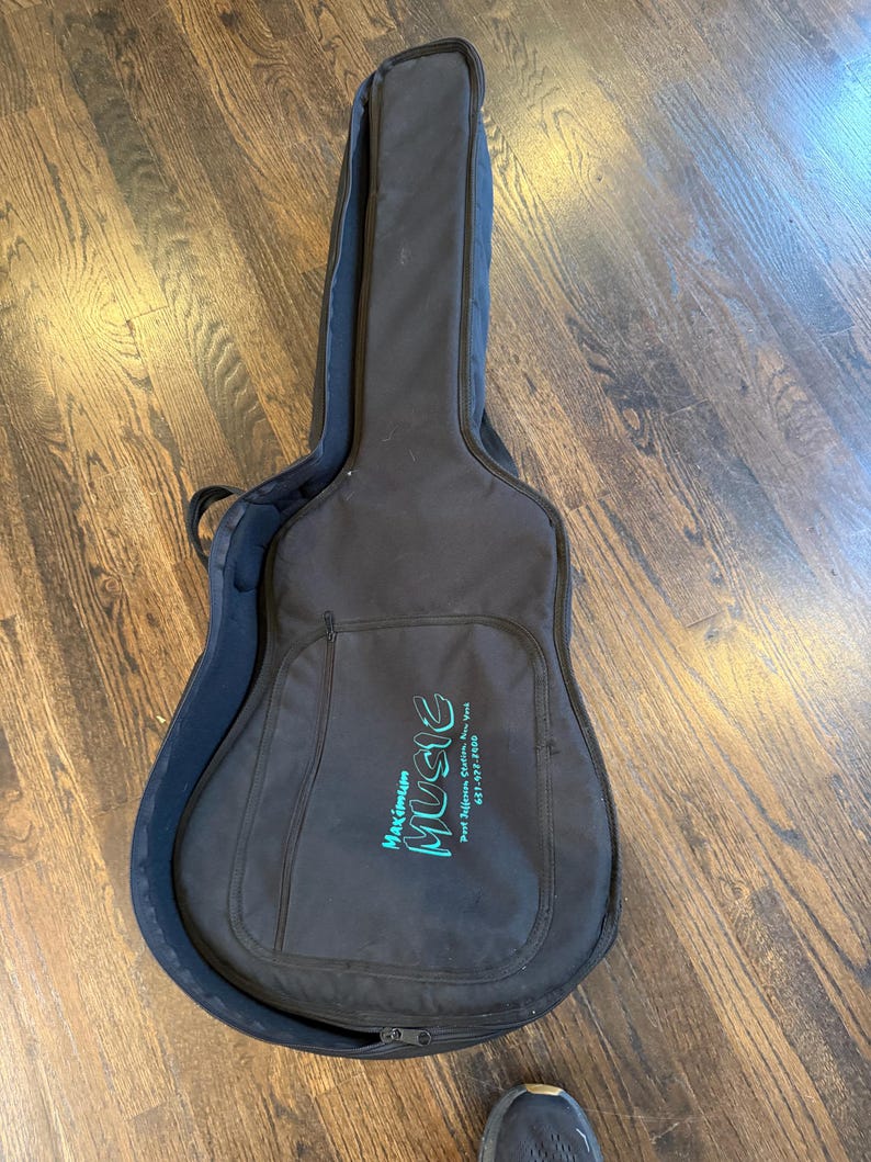 Pu&ograve; includere: Custodia per chitarra nera con chiusura a zip e tasca frontale. La custodia presenta la scritta "Maximum Music" in lettere turchesi. La custodia &egrave; aperta, rivelando un interno blu scuro. La custodia &egrave; su un pavimento in legno.