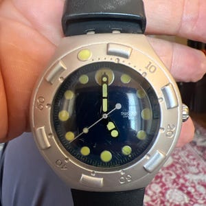 May include: A silver-toned Swatch watch with a black leather band. The watch face is dark blue with luminous green markers and hands. The bezel features numbers and a rotating design. The watch is a classic, sporty style.