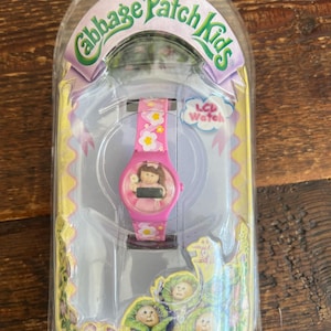 May include: A pink Cabbage Patch Kids LCD watch in its original packaging. The watch has a pink strap with white flower details and a round face with a Cabbage Patch Kids character. The packaging displays the Cabbage Patch Kids logo.
