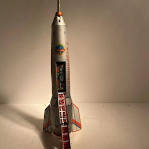 May include: A vintage silver toy rocket with red and orange accents, a ladder, and the text "HOLDRAKETA". The rocket has a detailed design, including a small astronaut figure inside.