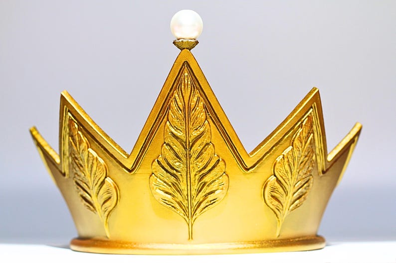May include: A gold-colored crown with a pearl at the top. The crown has a series of points and is decorated with leaf-like designs. The crown is likely a decorative item or part of a costume.