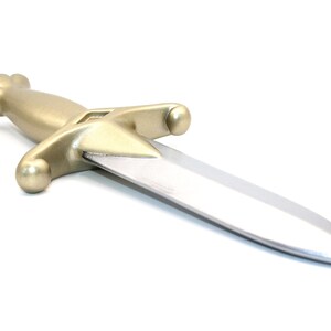 Könnte beinhalten: Ein Spielzeugdolch mit einem metallisch goldfarbenen Griff mit abgerundetem Knauf und Parierstange, kombiniert mit einer glatten, silberfarbenen Klinge, die zu einer abgerundeten Spitze zuläuft. Das Objekt ist schräg vor weißem Hintergrund.