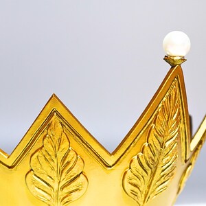 May include: A gold-colored crown with a pearl-like orb at the top. The crown has a leaf design and a pointed shape. The crown is likely made of metal and is a decorative item.