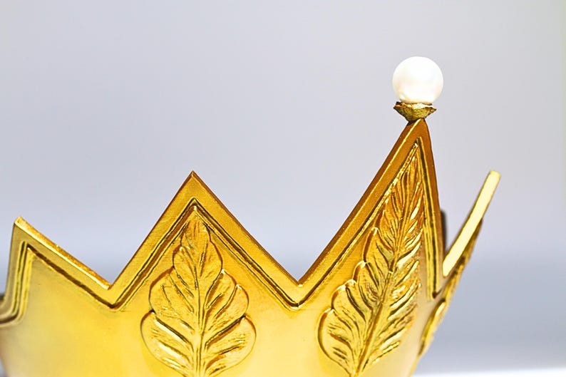 May include: A gold-colored crown with a pearl-like orb at the top. The crown has a leaf design and a pointed shape. The crown is likely made of metal and is a decorative item.