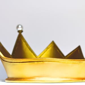 May include: A gold-colored crown with a pearl-like embellishment at the top. The crown has a series of triangular points and a rounded base. The crown is likely made of metal or a similar material.