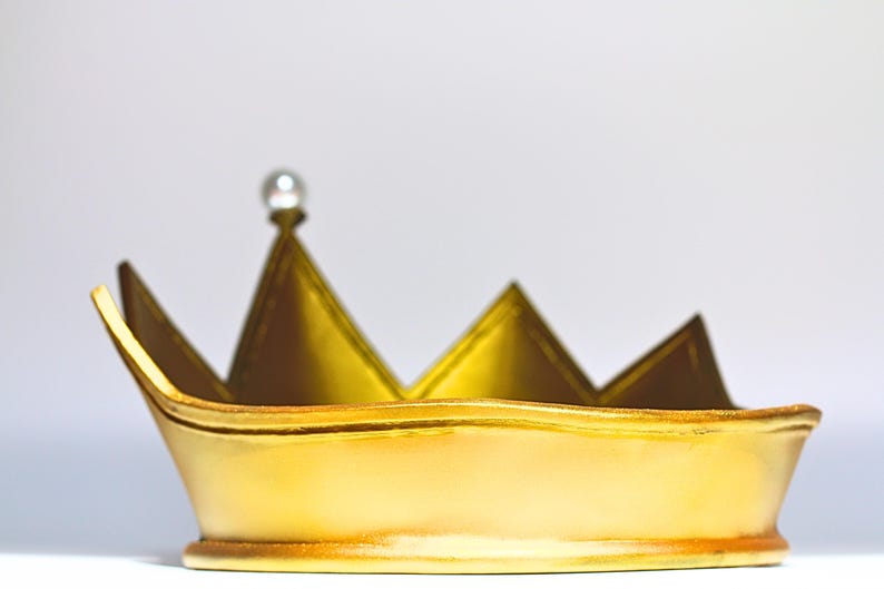 May include: A gold-colored crown with a pearl-like embellishment at the top. The crown has a series of triangular points and a rounded base. The crown is likely made of metal or a similar material.
