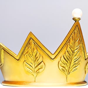 May include: A gold-colored crown with a pearl-like orb at the top. The crown features a zig-zag design with leaf-like embellishments. The crown is likely a decorative item or part of a costume.