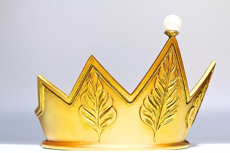 May include: A gold-colored crown with a pearl-like orb at the top. The crown features a zig-zag design with leaf-like embellishments. The crown is likely a decorative item or part of a costume.