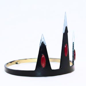 Può includere: Una corona nera con una base circolare e tre punte rivolte verso l'alto. Ogni punta è dotata di un'estremità color argento ed è decorata con una gemma ovale di colore rosso scuro incastonata al centro della struttura nera.