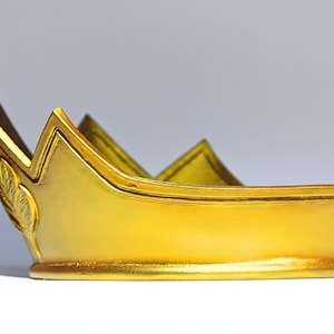 May include: A gold-colored crown with a pearl-topped spire. The crown has a rounded base and a series of upward-pointing peaks. A decorative leaf design is visible on the side. The crown is likely a costume accessory or decorative item.