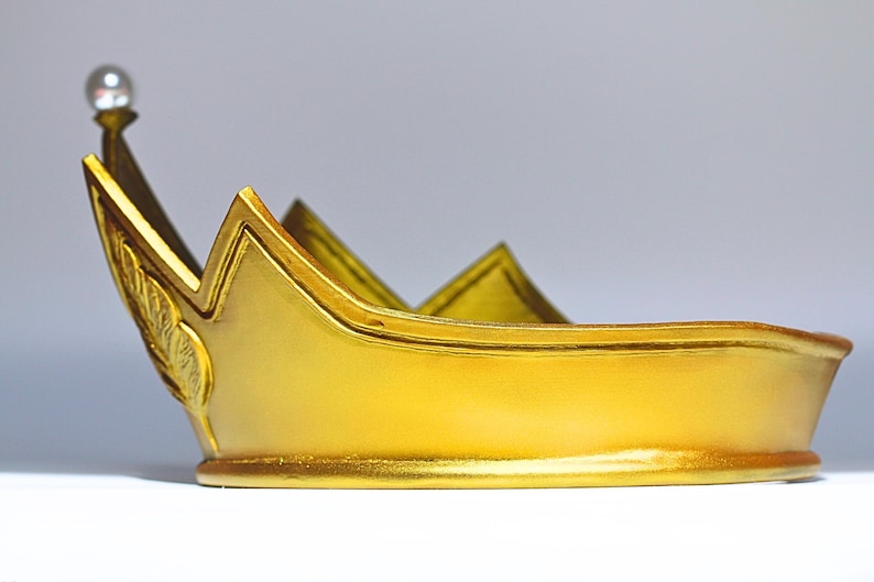 May include: A gold-colored crown with a pearl-topped spire. The crown has a rounded base and a series of upward-pointing peaks. A decorative leaf design is visible on the side. The crown is likely a costume accessory or decorative item.