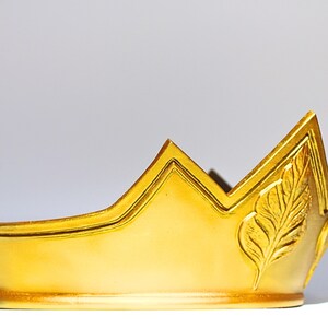 May include: A gold-colored crown with a pearl-topped spire. The crown features a leaf design and a pointed peak. The crown is likely made of metal and is designed to be worn. The crown is a symbol of royalty and is often used in costume or theatrical settings.