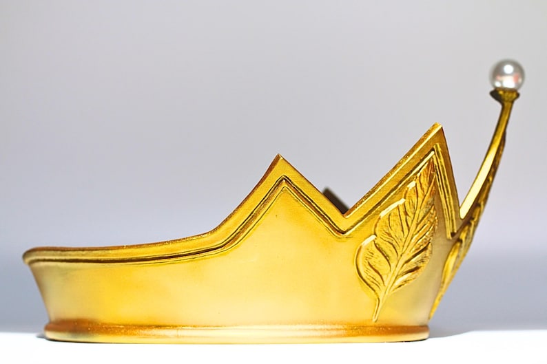 May include: A gold-colored crown with a pearl-topped spire. The crown features a leaf design and a pointed peak. The crown is likely made of metal and is designed to be worn. The crown is a symbol of royalty and is often used in costume or theatrical settings.