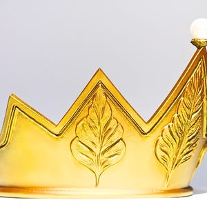 May include: A gold-colored crown with a pearl-like orb at the highest point. The crown features leaf-like designs and a zig-zag top. The crown is likely a decorative item, possibly for a costume or display.