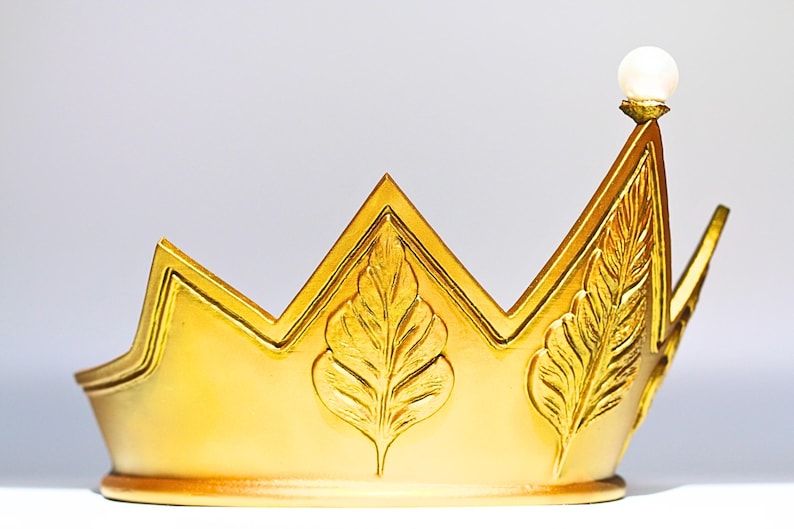May include: A gold-colored crown with a pearl-like orb at the highest point. The crown features leaf-like designs and a zig-zag top. The crown is likely a decorative item, possibly for a costume or display.