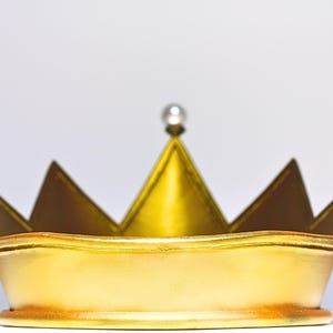 May include: A gold-colored crown with a pearl-like bead at the top. The crown has a classic design with pointed tips and a rounded base. The crown is likely made of metal and has a shiny finish.