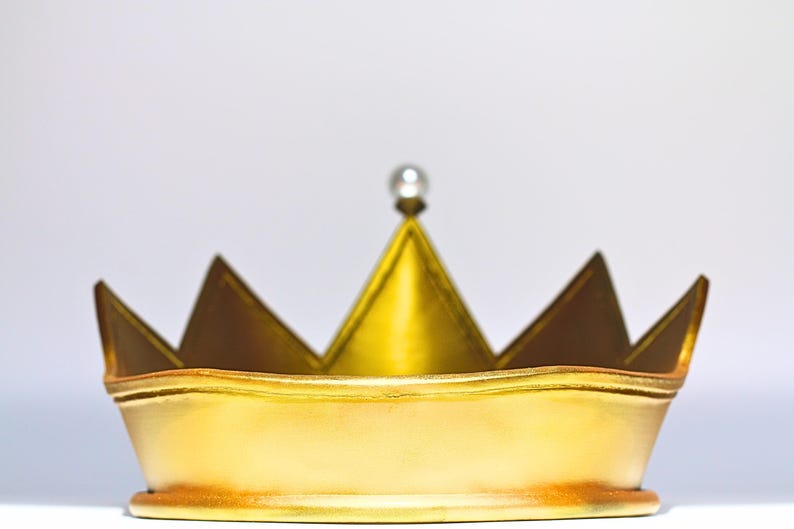 May include: A gold-colored crown with a pearl-like bead at the top. The crown has a classic design with pointed tips and a rounded base. The crown is likely made of metal and has a shiny finish.