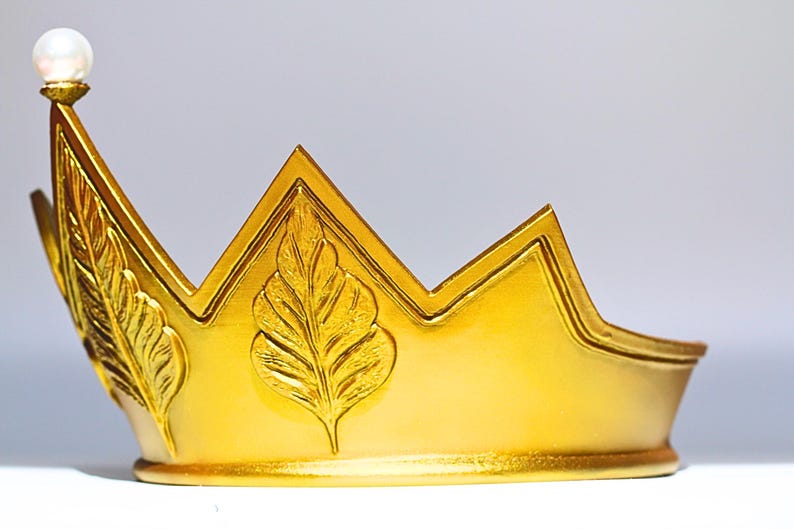 May include: A golden crown with a pearl at the top. The crown has a series of points and is decorated with leaf designs. The crown is made of a shiny gold material and is likely a decorative item.