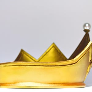 May include: A gold-colored crown with a pearl at the top. The crown has a rounded base and pointed peaks. The crown is made of a shiny material and appears to be a costume piece or decorative item.