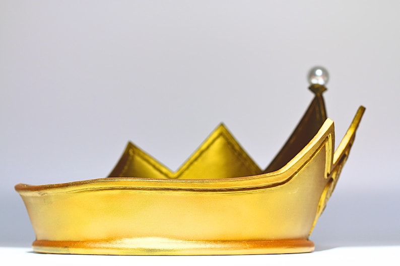 May include: A gold-colored crown with a pearl at the top. The crown has a rounded base and pointed peaks. The crown is made of a shiny material and appears to be a costume piece or decorative item.