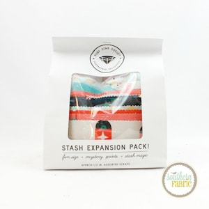 May include: A white paper bag labeled "STASH EXPANSION PACK!" containing assorted fabric scraps. The bag features a clear window revealing colorful, patterned fabric strips. The text includes "fun size," "mystery prints," and "stash magic."