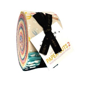 Peut inclure: Un rouleau de bandes de tissu avec divers motifs colorés. Le paquet est noué avec un ruban noir et une étiquette indiquant "PAPER CUTS 2" et "RASHIDA COLEMAN HALE". Le tissu a un fond crème avec des couleurs pastel.