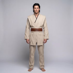 May include: A beige Jedi-style costume with a brown leather belt. The outfit includes a long-sleeved tunic with a dark brown trim, matching trousers, and a wide belt with a metal buckle. The costume is designed for cosplay or themed events.