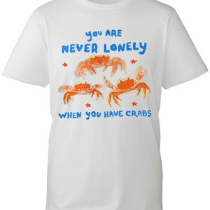 May include: A white short-sleeved t-shirt featuring a graphic of three orange crabs and small red stars. The blue text reads, "YOU ARE NEVER LONELY WHEN YOU HAVE CRABS," positioned above and below the illustrated crab design on the front.